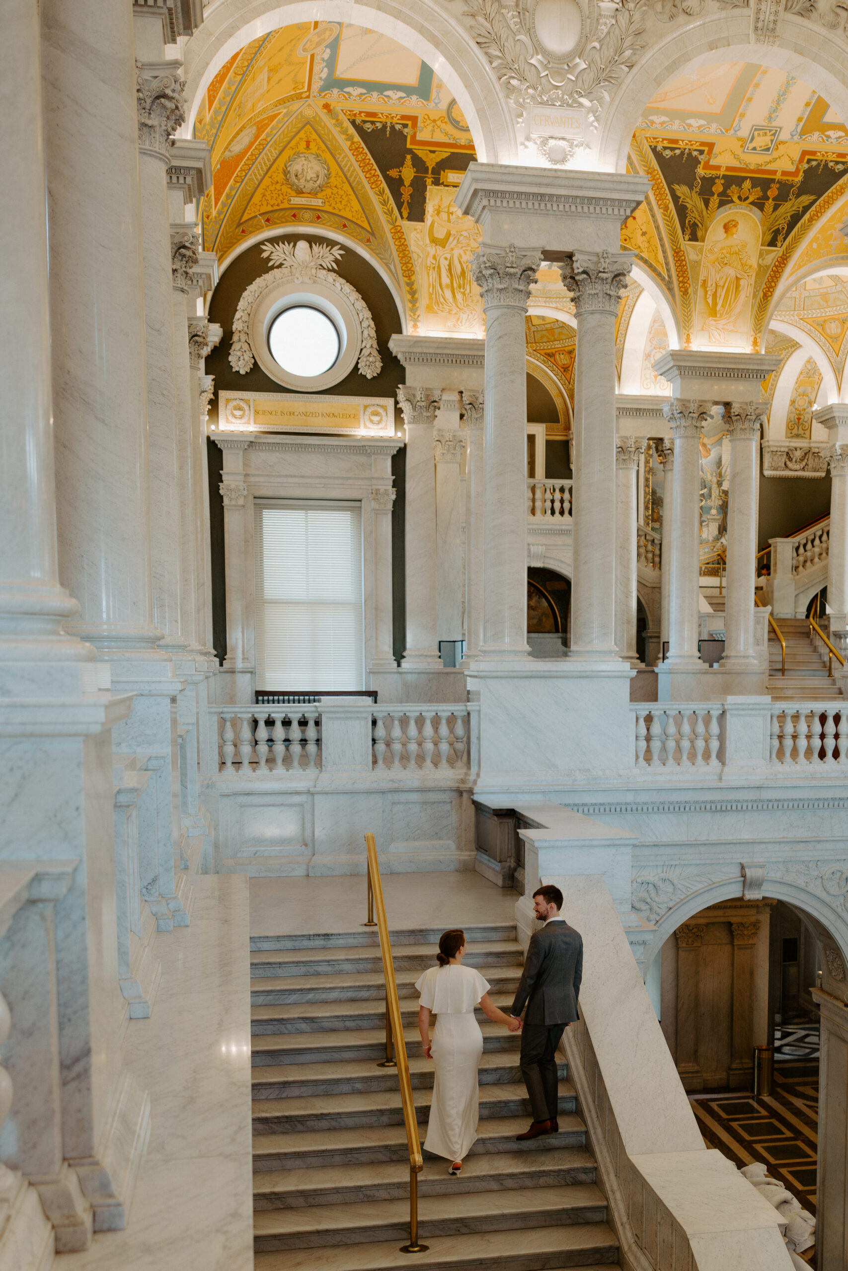 Wide shot of a couple holding hands on the grand staircase of the Library of Congress during their DC engagement session, surrounded by ornate marble architecture