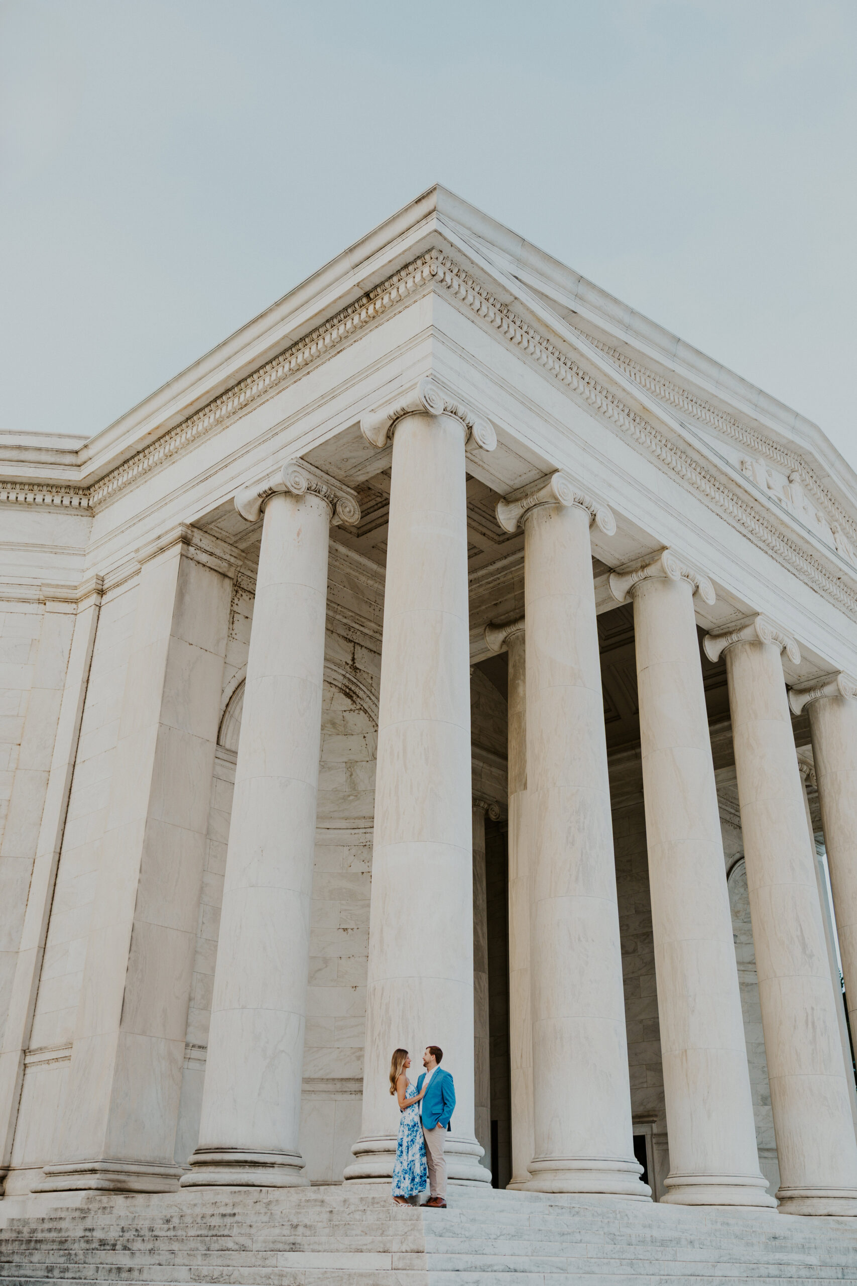 Wide shot of the Jefferson Memorial’s grand architecture with a couple standing at the base of the columns for their engagement photos.